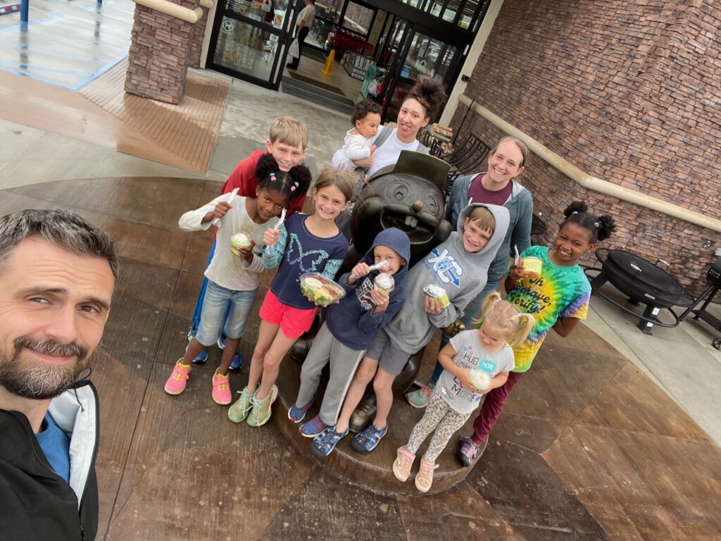 Group of people posing for a selfie with a statue, some holding ice cream cones, in an outdoor shopping area.